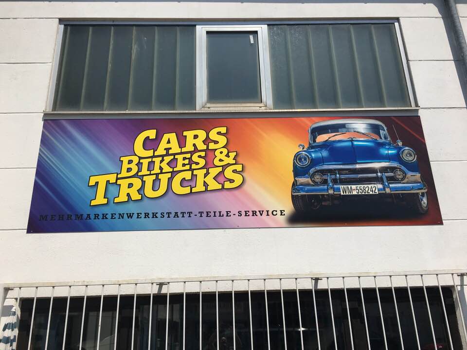 Buntes Werbeschild mit der Aufschrift 'Cars, Bikes & Trucks' und einem klassischen blauen Auto auf der linken Seite.